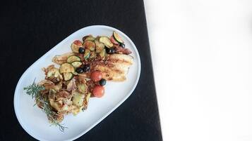 Une assiette ovale blanche contenant un plat composé de poisson grillé, de pommes de terre rôties, de rondelles de courgettes, de tomates cerises et d’olives noires, le tout agrémenté d’herbes aromatiques.