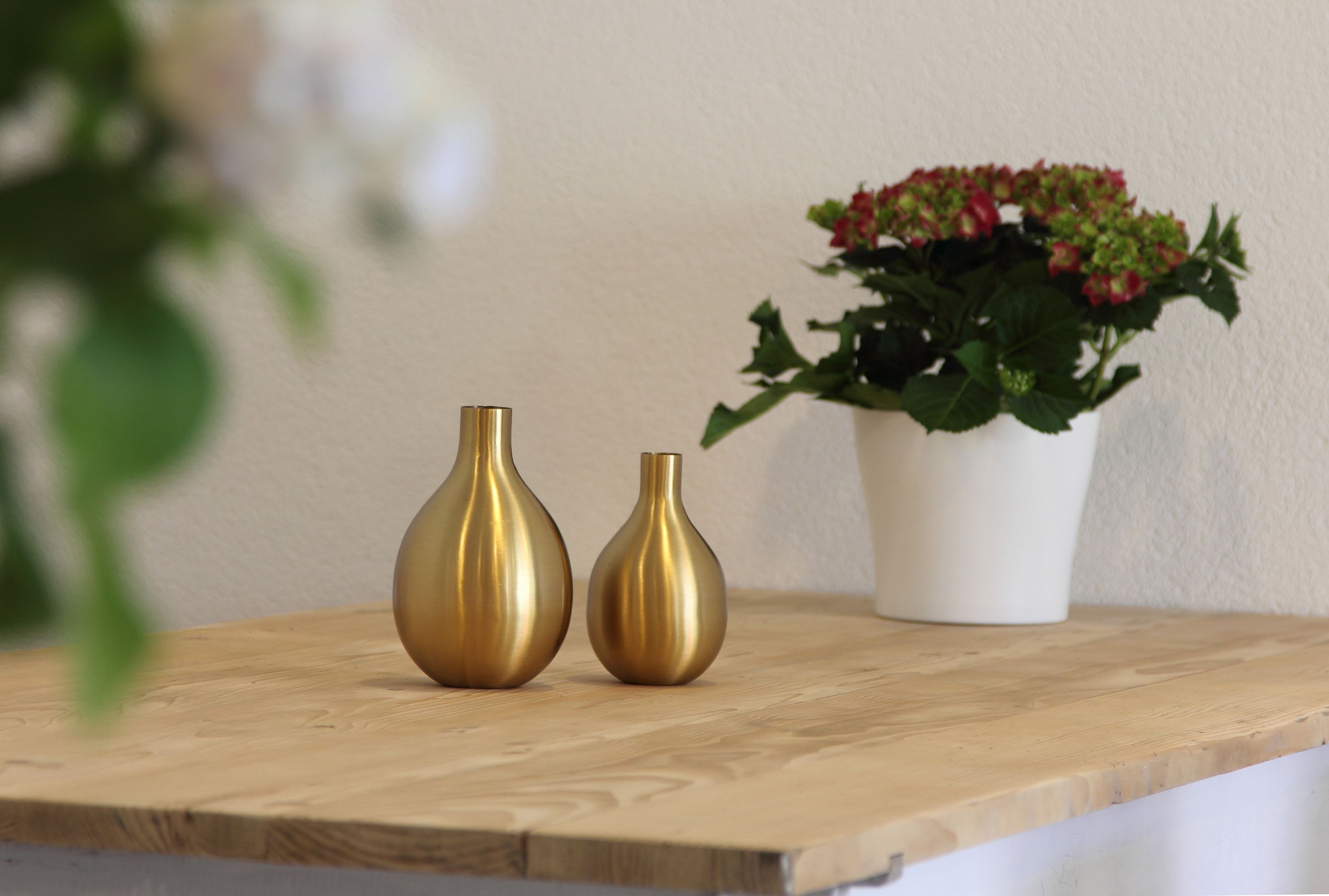 Deux vases en or sont posés sur une surface en bois, à côté d’un pot blanc contenant des fleurs rouges.