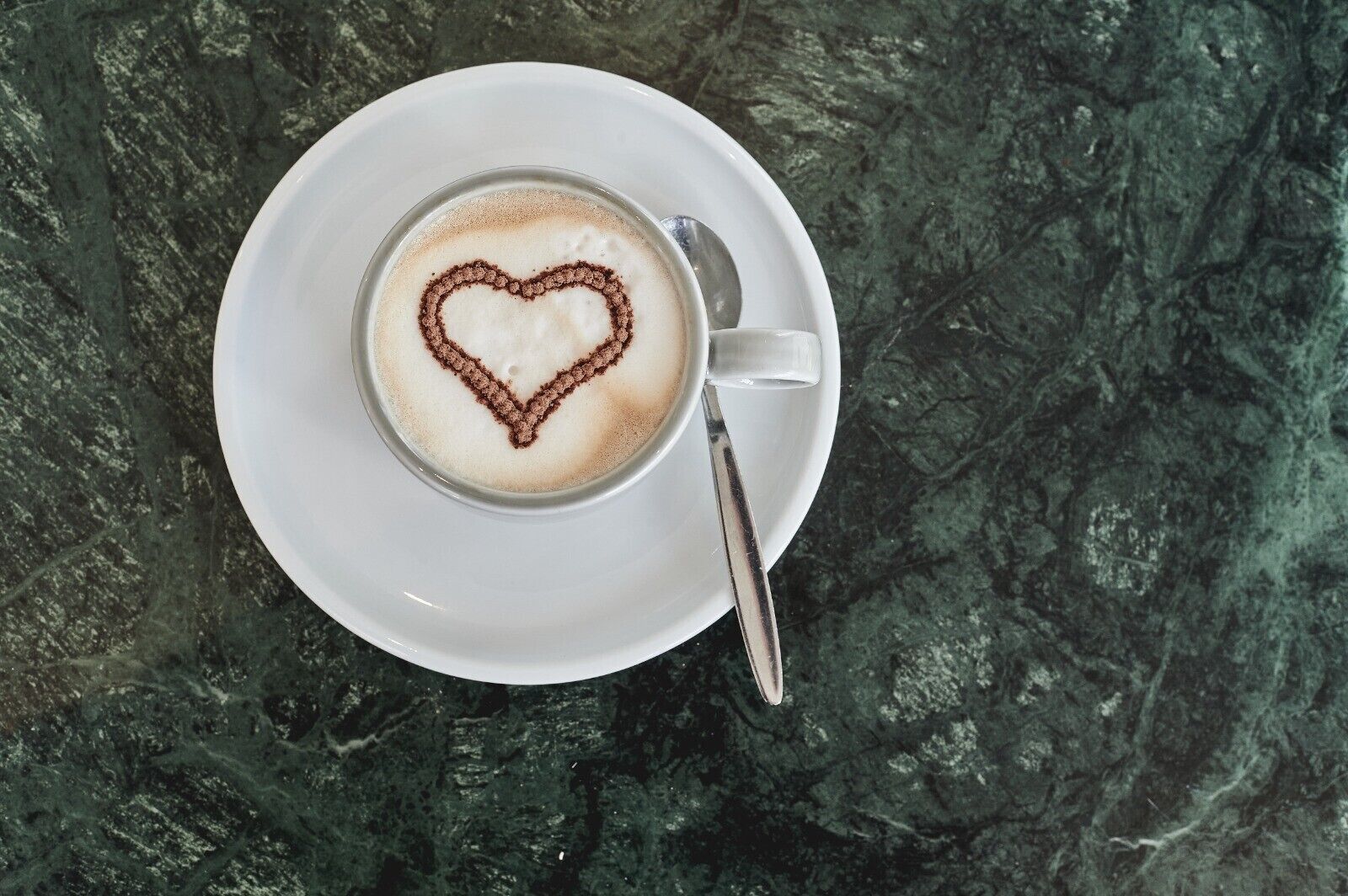 A cup of coffee with a heart-shaped latte art on a white saucer, placed on a dark marble surface.