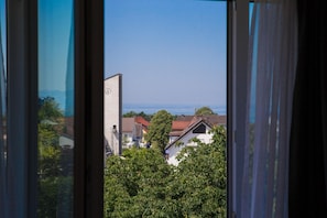 A view through a window with curtains, featuring a building with a clock, surrounded by greenery and houses. - Adler Hotel & Gasthaus (Nonnenhorn)