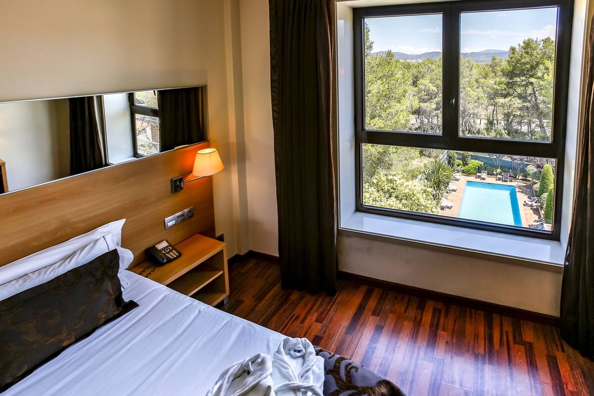 Quarto de hotel com cama, mesa de cabeceira, abajur e uma grande janela com vista para uma piscina e árvores.