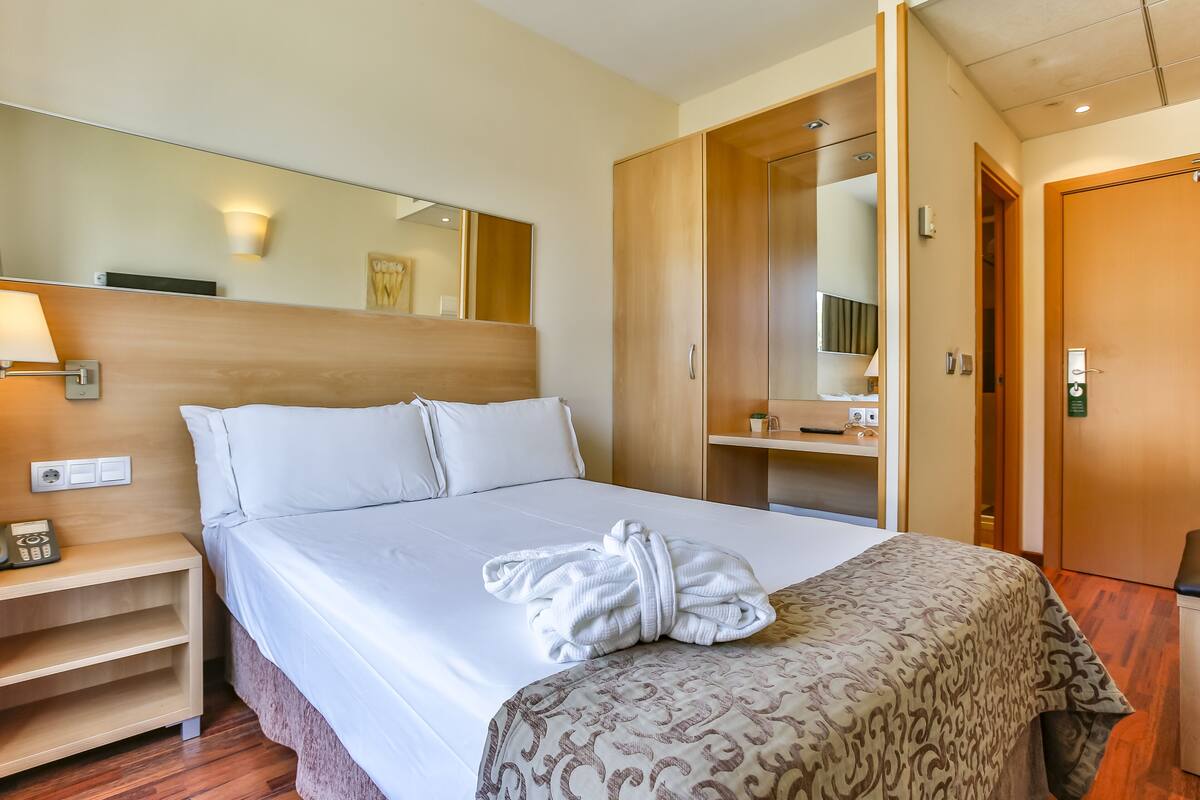 Quarto de hotel com cama bem arrumada, mesa de cabeceira com abajur, roupão sobre a cama e guarda-roupa com espelho.