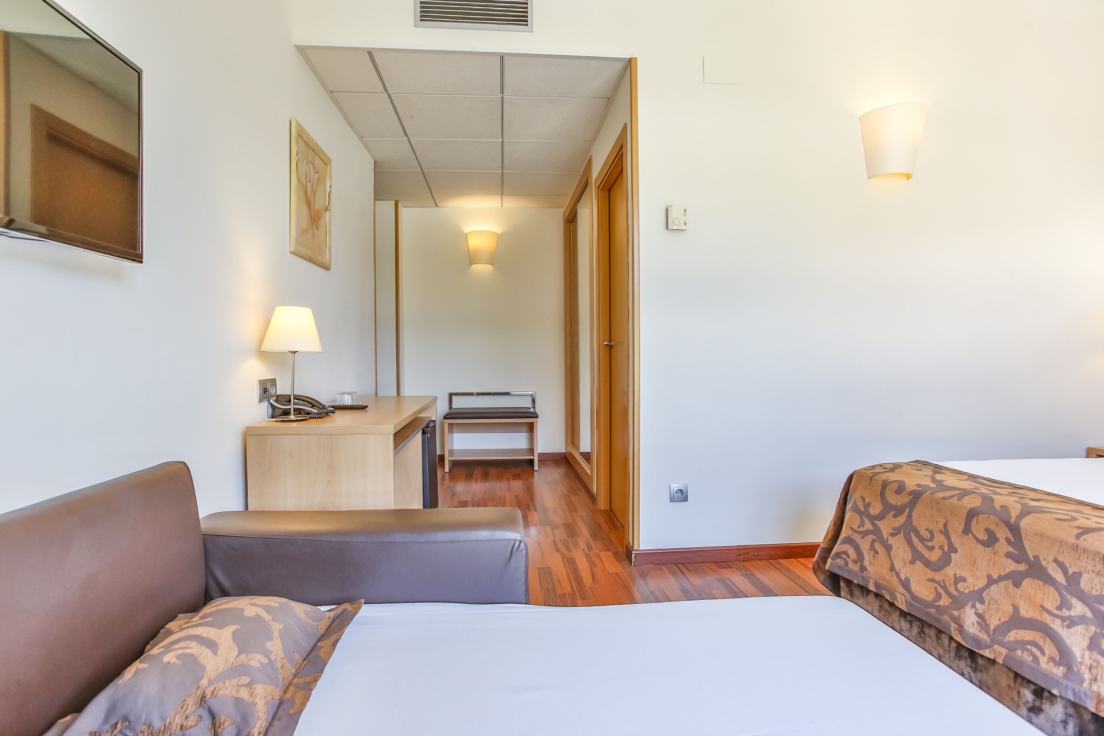 A hotel room with a bed, a desk with a lamp, a chair, and a wooden floor.