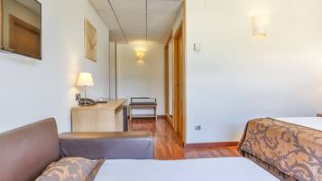 A hotel room with a bed, a desk with a lamp, a chair, and a wooden floor.