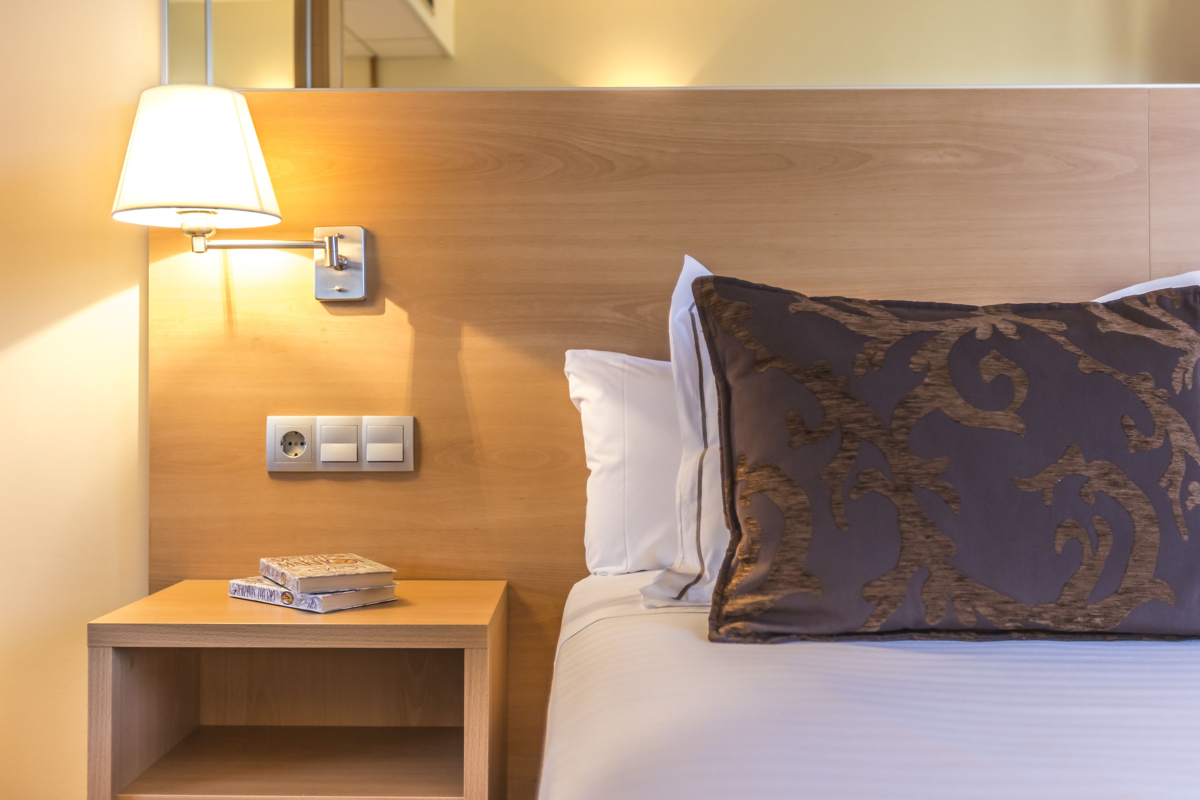 Una habitación de hotel con cabecera de madera, lámpara de noche, mesita de noche con libros y un cojín decorativo.