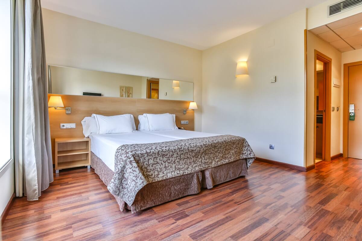 Quarto de hotel com uma cama grande, piso de madeira, um espelho sobre a cama e uma mesa de cabeceira com uma luminária.