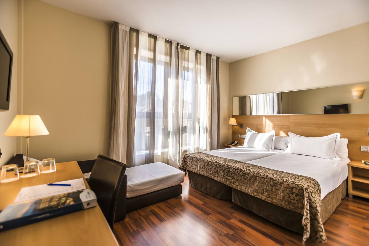 Quarto de hotel com cama, escrivaninha com luminária, cadeira, mesa de cabeceira e uma grande janela com cortinas.