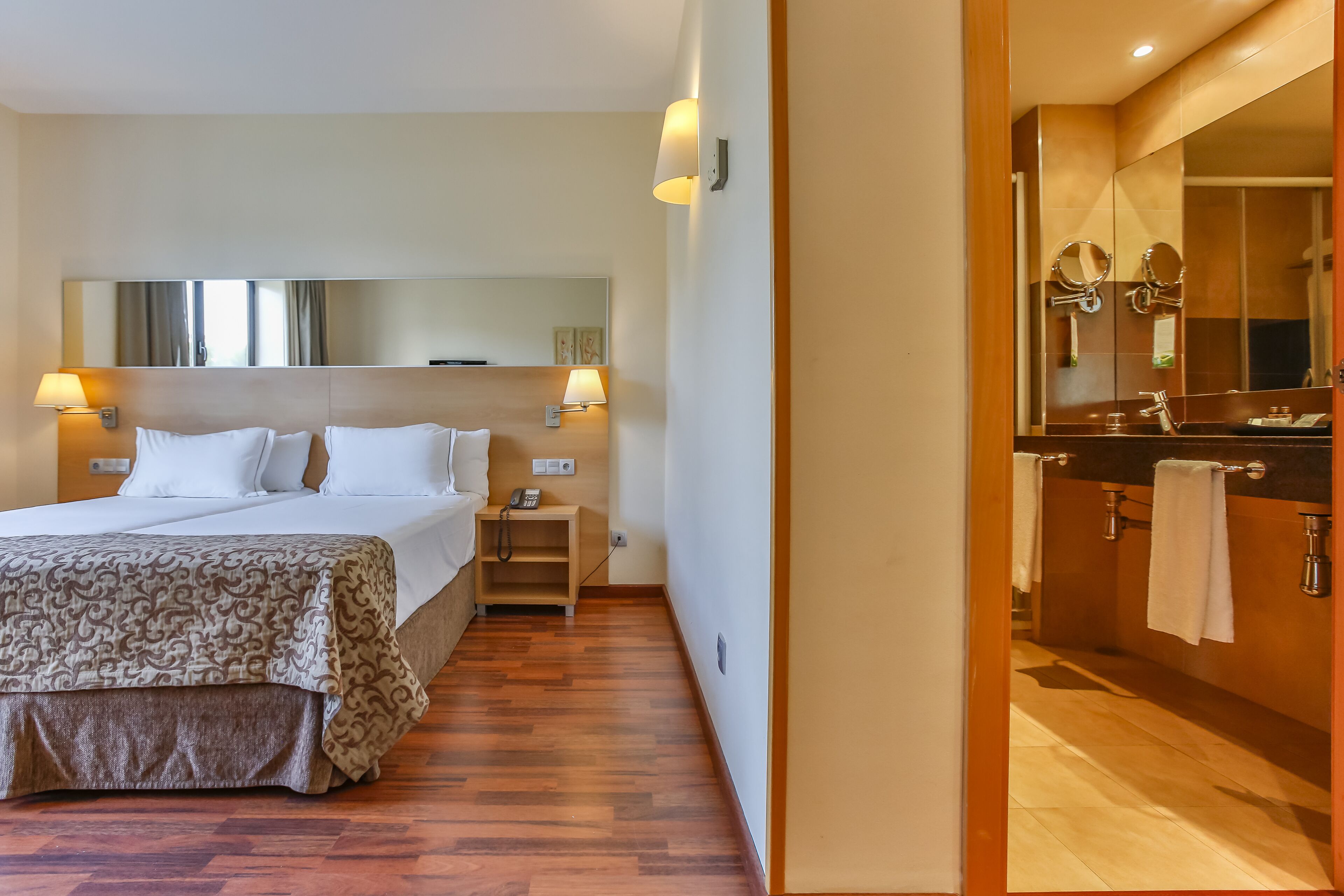 Habitación de hotel con cama, lámparas de noche, mesita de noche, espejo y baño con lavamanos y perchero para toallas.