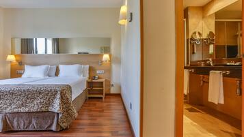Habitación de hotel con cama, lámparas de noche, mesita de noche, espejo y baño con lavamanos y perchero para toallas.