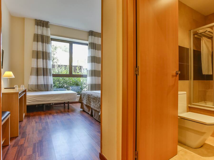 Una habitación pequeña con cama, una mesita con lámpara, ventana con cortinas y baño con inodoro y ducha.