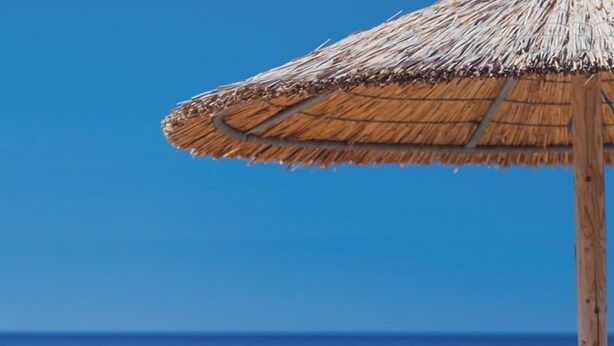 Two wooden lounge chairs with a thatched umbrella, a person sitting under the umbrella, and a clear view of the ocean. - Cavo Petra (Troizinia)