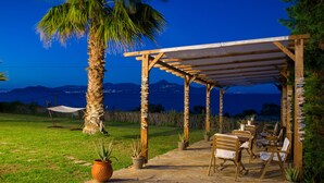 A covered outdoor seating area with chairs and tables, surrounded by potted plants and a palm tree. - Cavo Petra (Troizinia)
