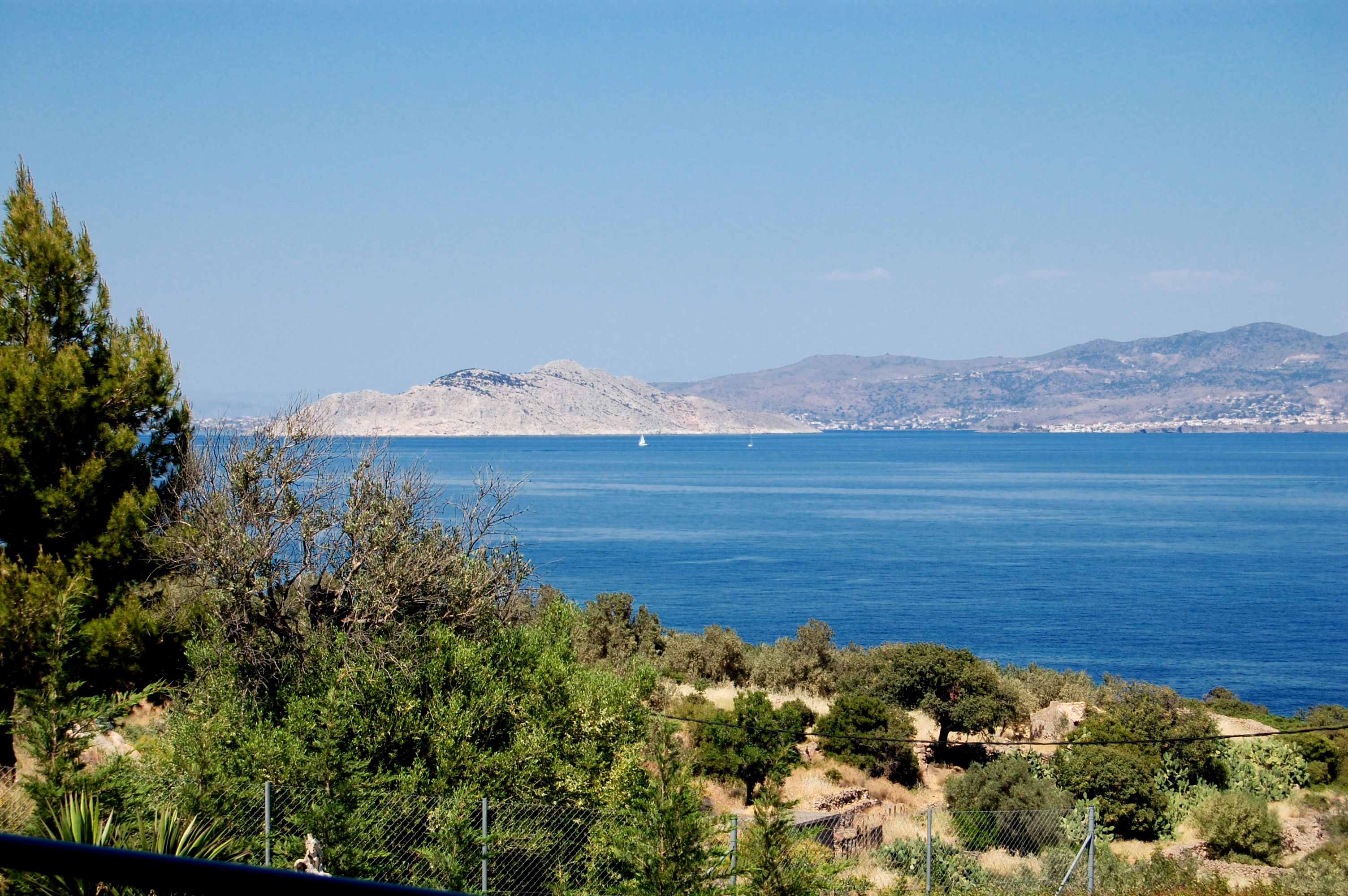 A coastal landscape with a clear view of the sea, a distant mountain, and a variety of vegetation including trees and shrubs.