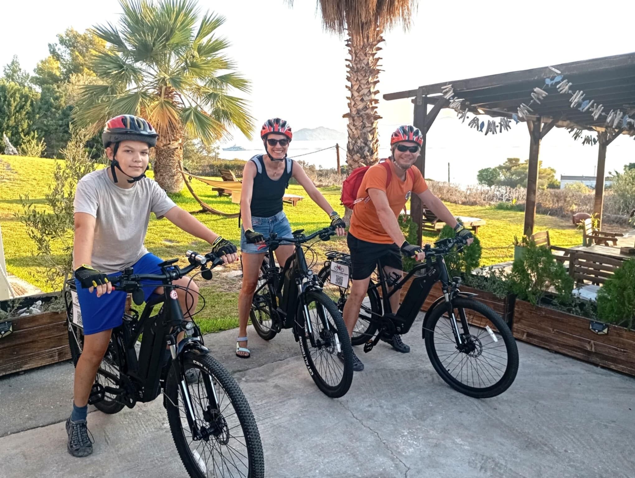 Três pessoas em bicicletas em ambiente externo, com uma estrutura de madeira e vegetação.