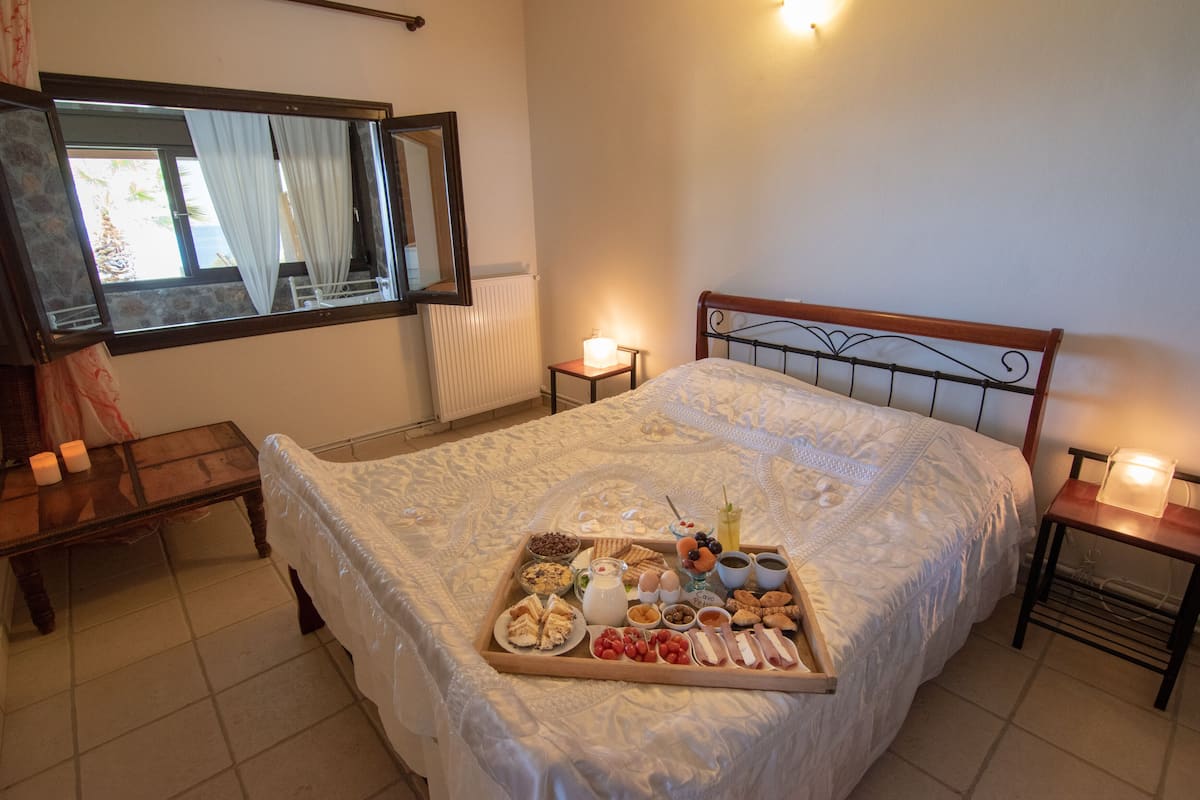 A neatly made bed with a tray of breakfast items, a bedside table with a lamp, and a window with curtains.