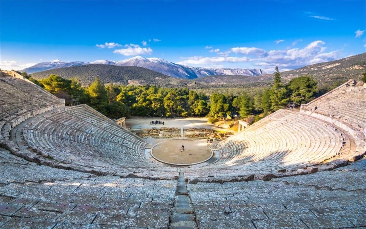 Un anfiteatro antiguo con asientos escalonados y un escenario central, rodeado de árboles y montañas al fondo.