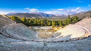Un anfiteatro antiguo con asientos escalonados y un escenario central, rodeado de árboles y montañas al fondo.