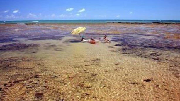A beach with clear shallow waters, a person lying on the sand, and another person sitting under an umbrella. - Villa Flamboyan (Lauro de Freitas)