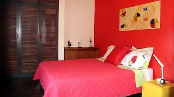 Quarto com colcha vermelha, cabeceira de madeira, mesa de cabeceira com luminária e um quadro na parede.