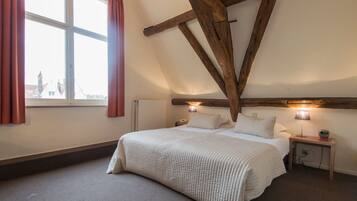 A bedroom with a sloped ceiling, a bed with white bedding, two bedside lamps, a nightstand, and a window with curtains.