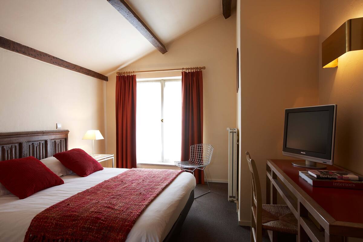 A hotel room with a bed, a desk with a computer, a chair, and a window with curtains.