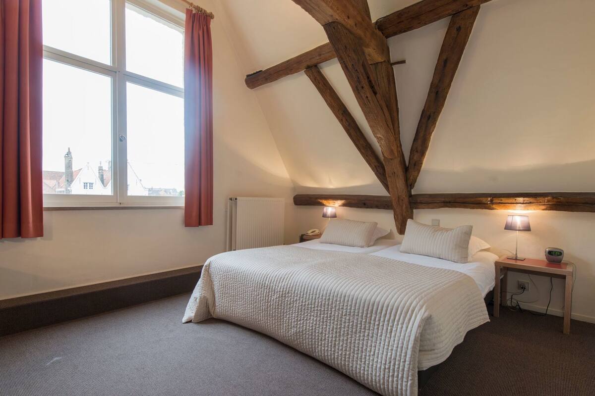 A bedroom with a large bed, two bedside lamps, a wooden ceiling beam, and a window with curtains.