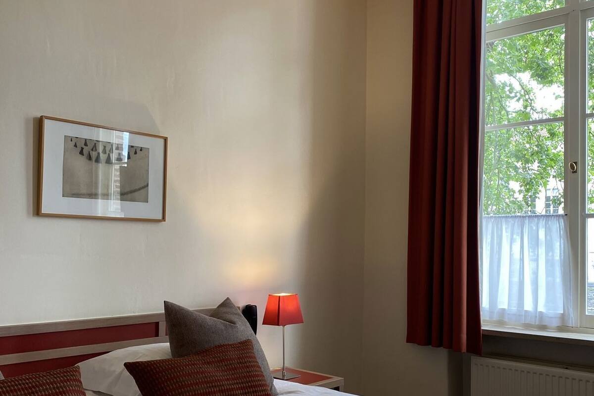 A hotel room with two beds, a framed picture on the wall, a window with curtains, and a nightstand with a lamp.