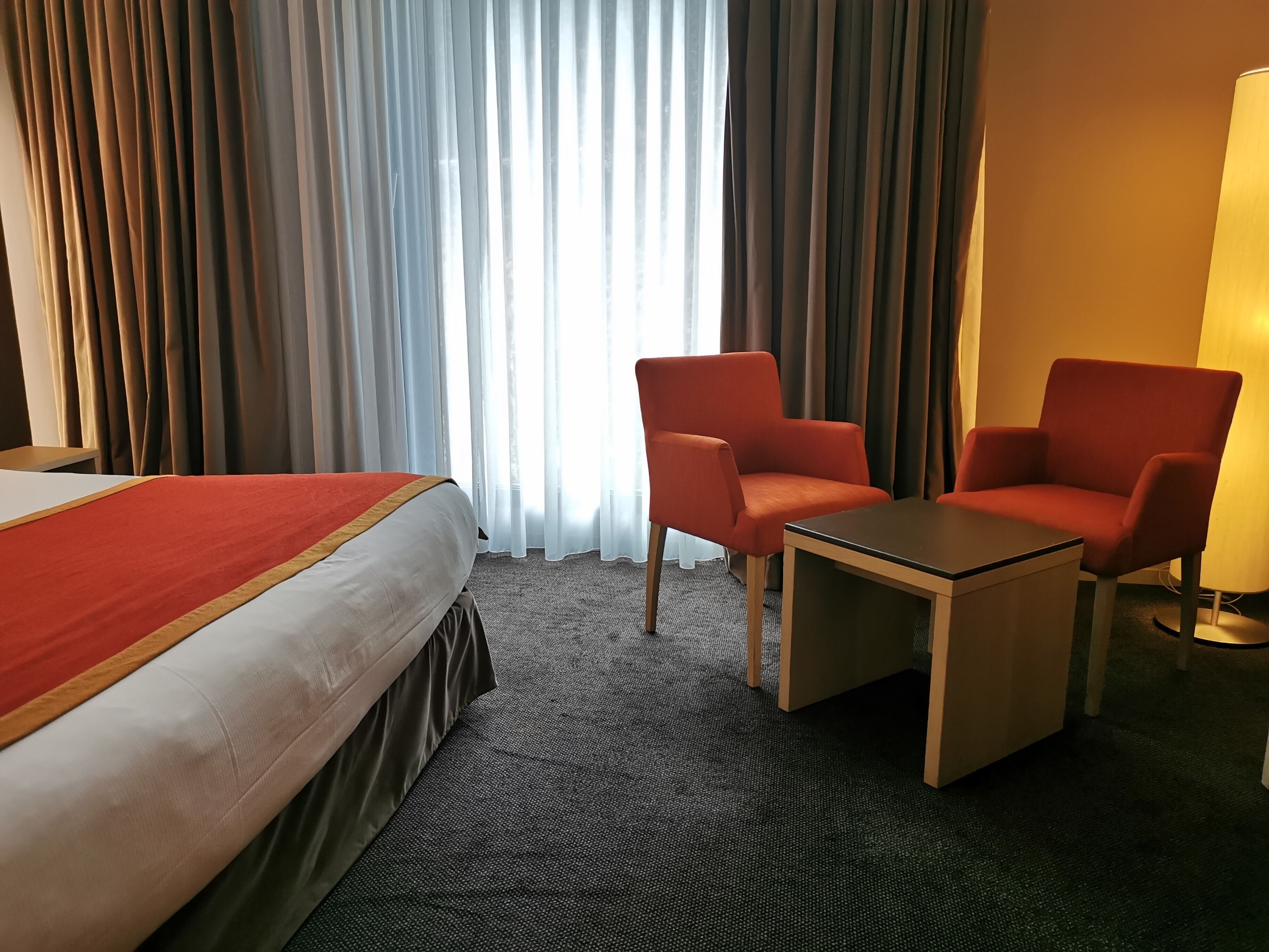 A hotel room with a bed, two red armchairs, a small table, and a lamp.