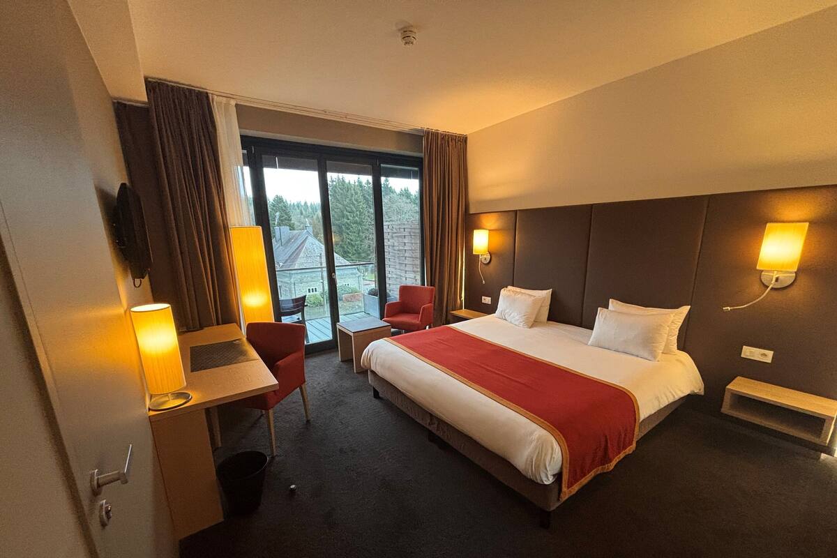 A hotel room with a large bed, a desk with a chair, a small table, and a window with curtains.