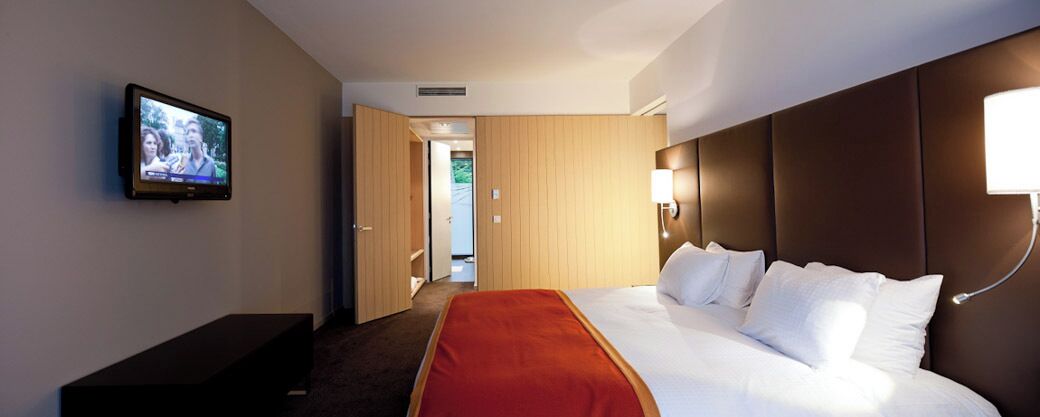 Une chambre d’hôtel avec un lit, un téléviseur fixé au mur et une porte donnant sur un couloir.