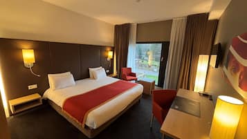 Een hotelkamer met een groot bed, een bureau met stoel, een lamp en uitzicht op het omliggende landschap door een raam.