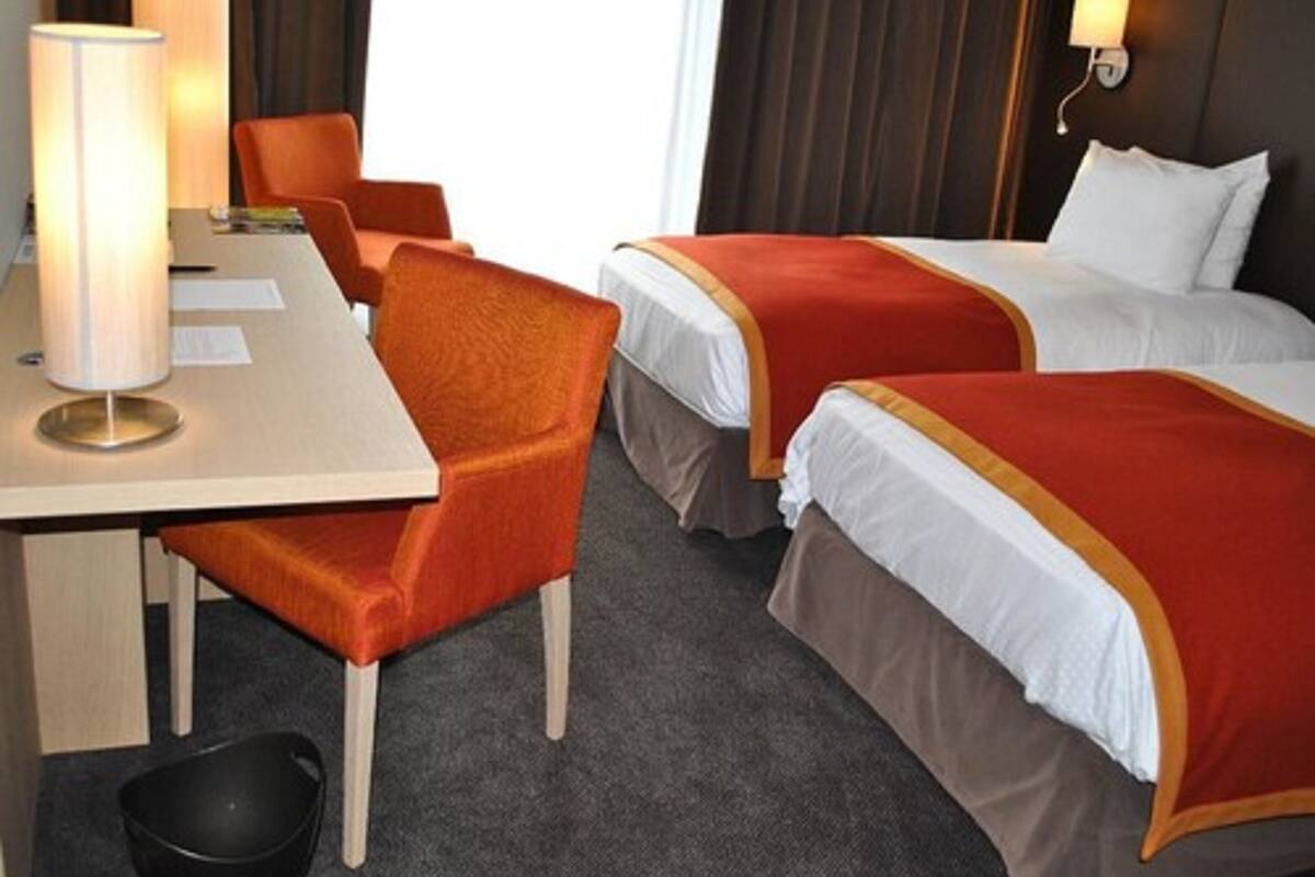 A hotel room with two beds, a desk, a chair, a lamp, and a wall-mounted light.