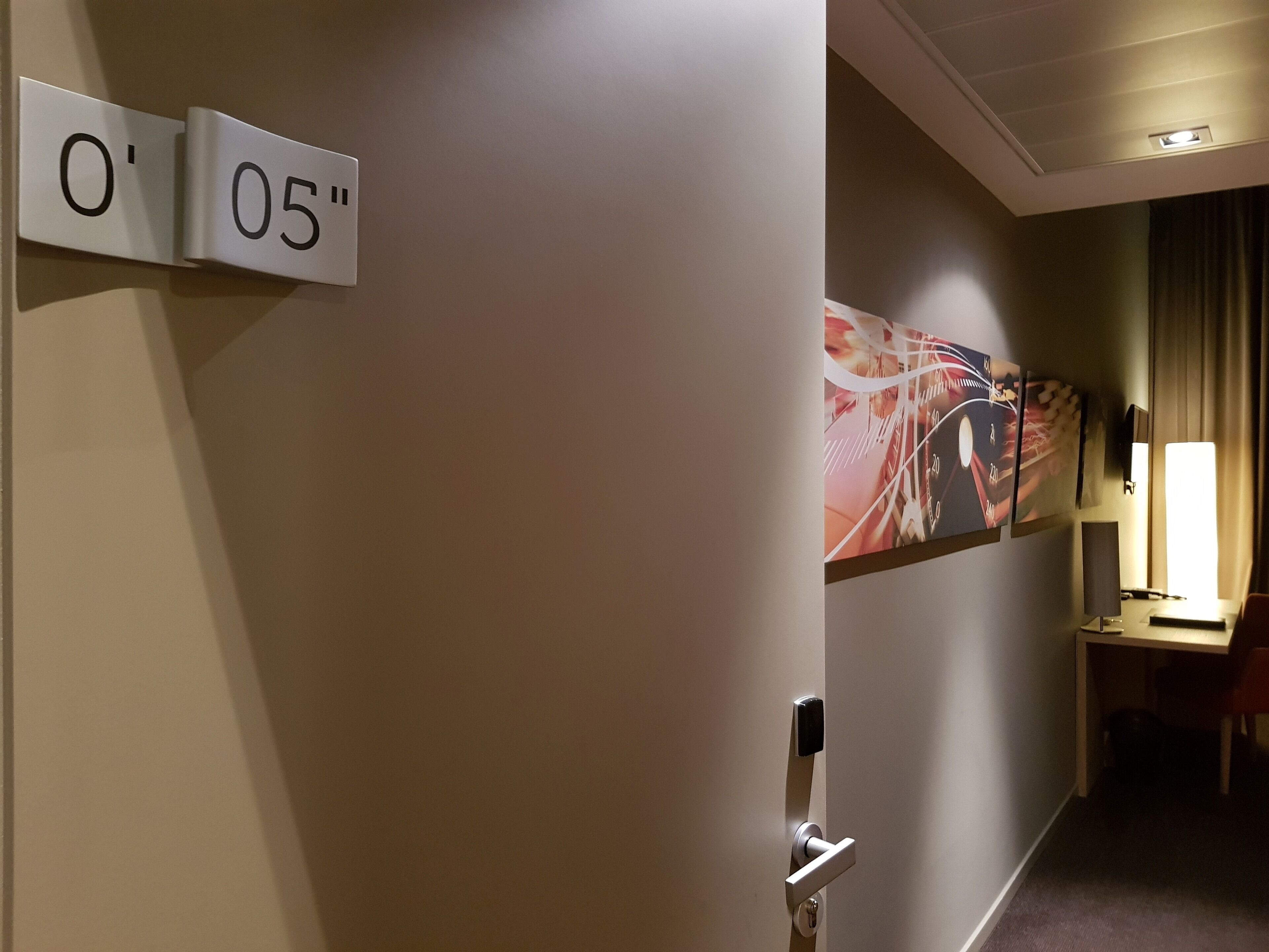 Un couloir d’hôtel avec une porte portant les numéros « 0 » et « 05 », un bureau avec une lampe et un tableau accroché au mur.