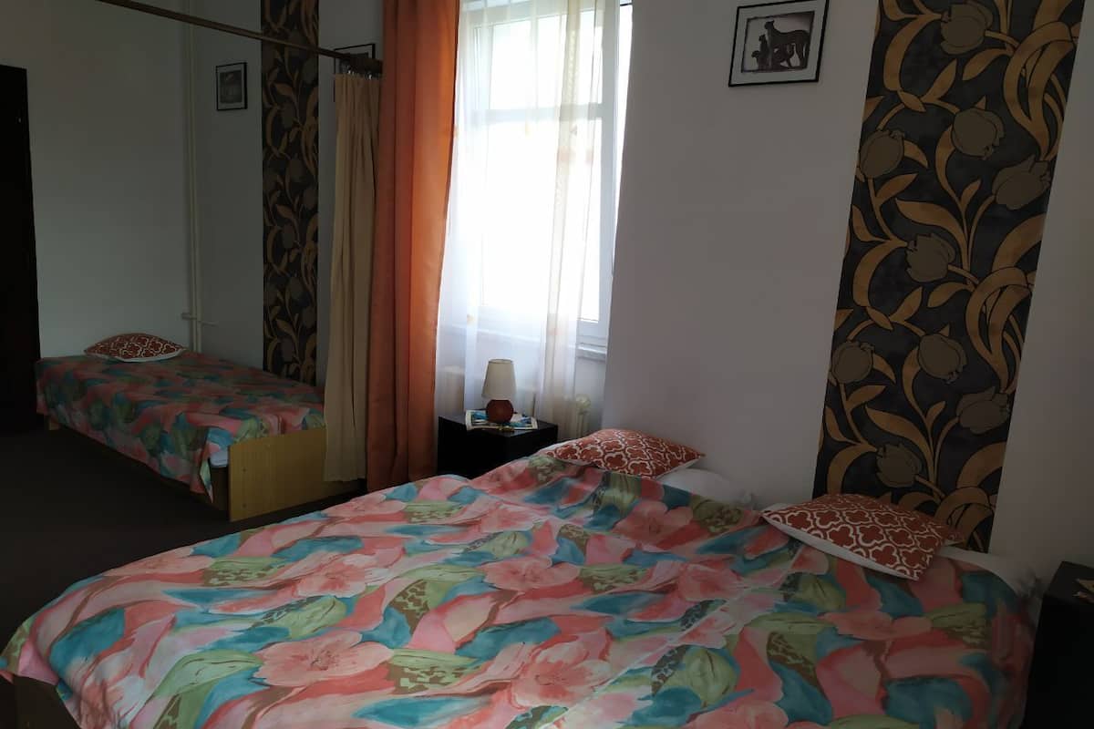 A bedroom with two beds, a window with sheer curtains, a bedside table with a lamp, and a floral-patterned wall.