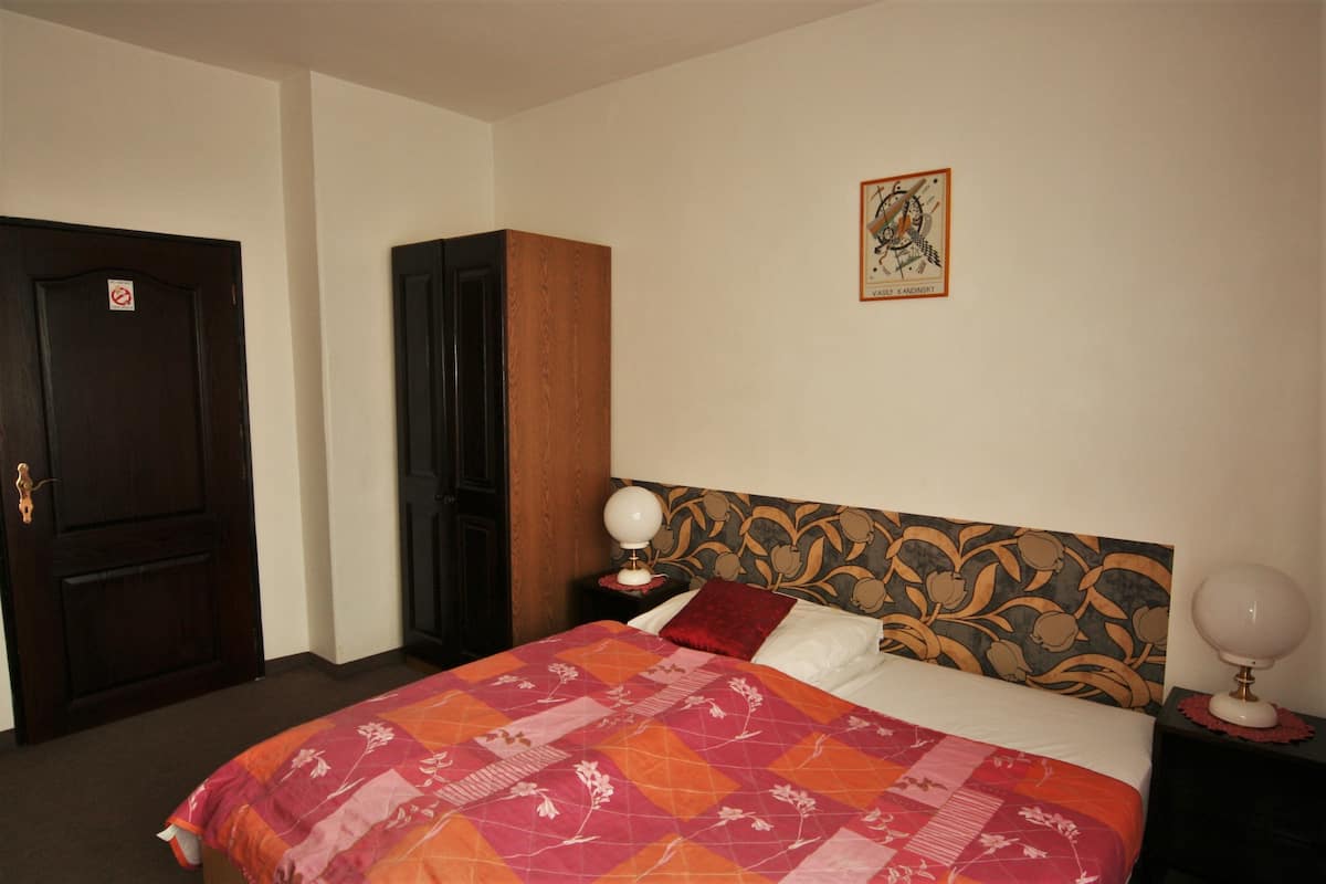A bedroom with a bed, a nightstand, a lamp, a door, and a wall with a framed picture.