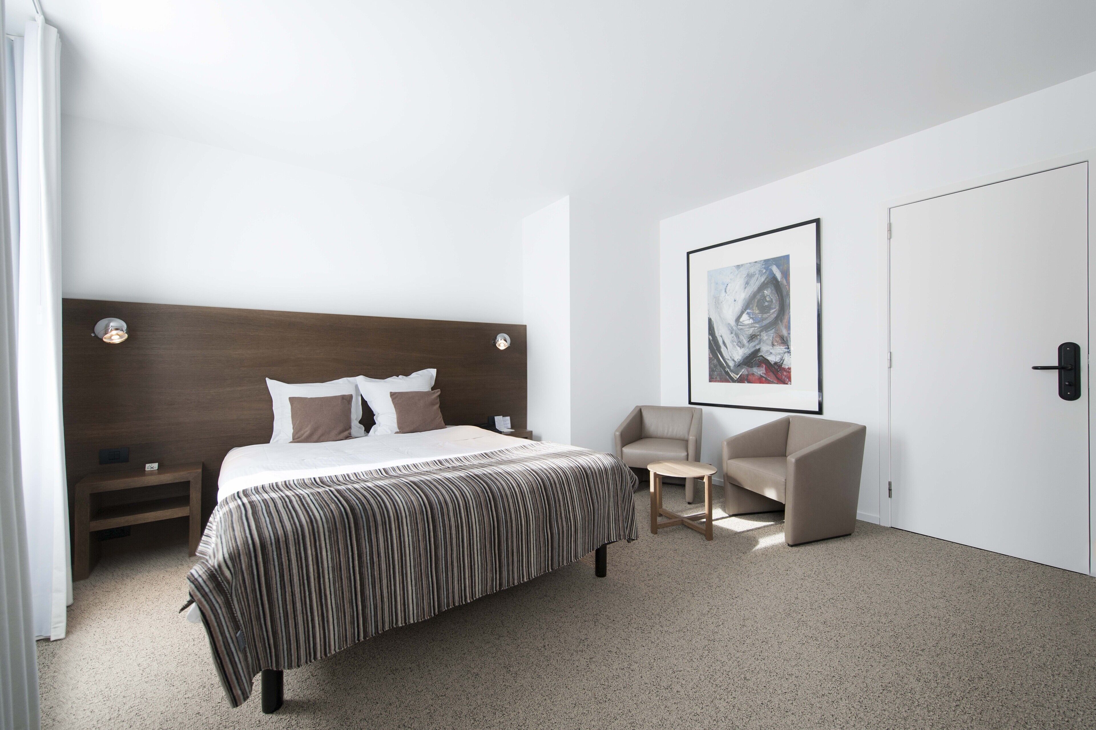 Ett modernt hotellrum med en stor säng, en sänggavel, ett nattduksbord, en stol, ett litet bord och ett inramat konstverk på väggen.