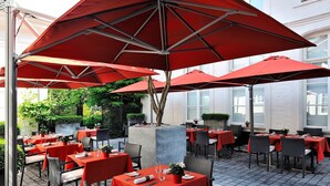 Un área de comedor al aire libre con sombrillas rojas y mesas, rodeada de follaje y un edificio al fondo.