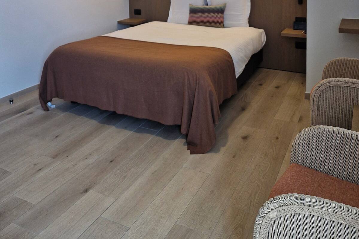 A modern hotel room with a large bed, a headboard with integrated lighting, a framed artwork on the wall, and a wicker chair.