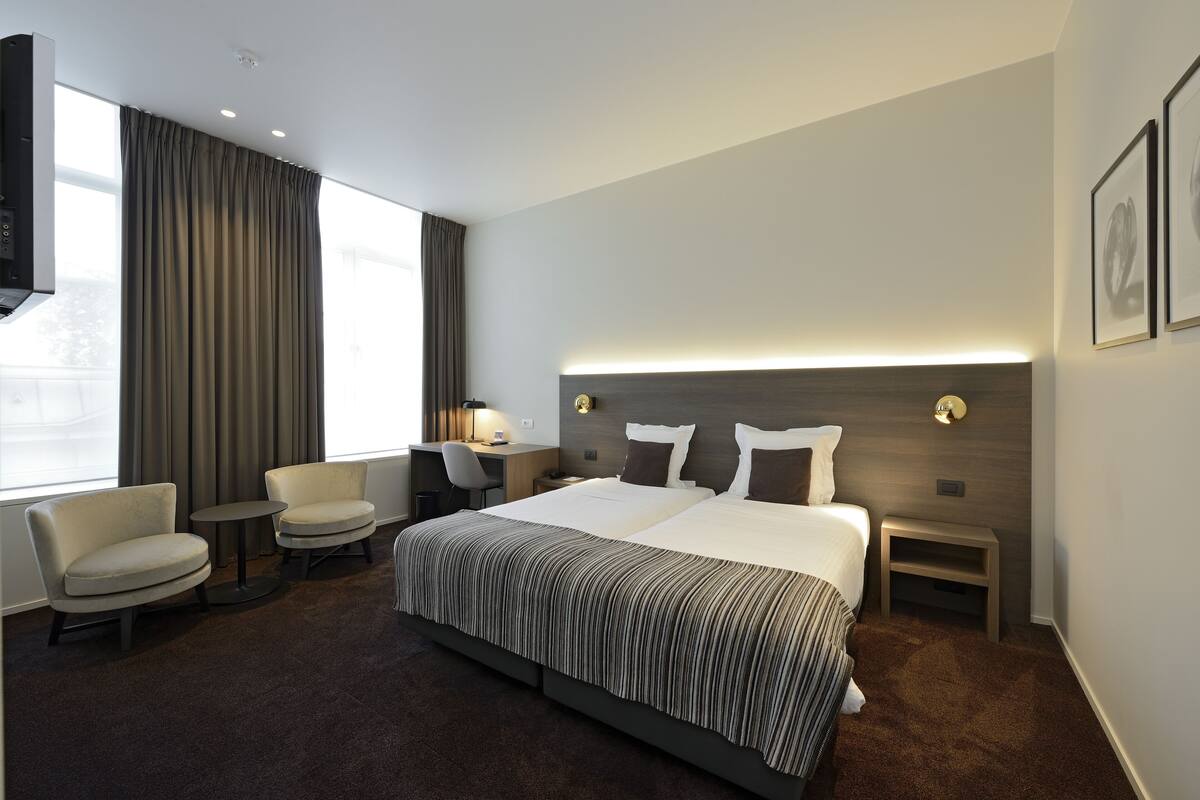 A modern hotel room with a large bed, two armchairs, a desk, and a TV.