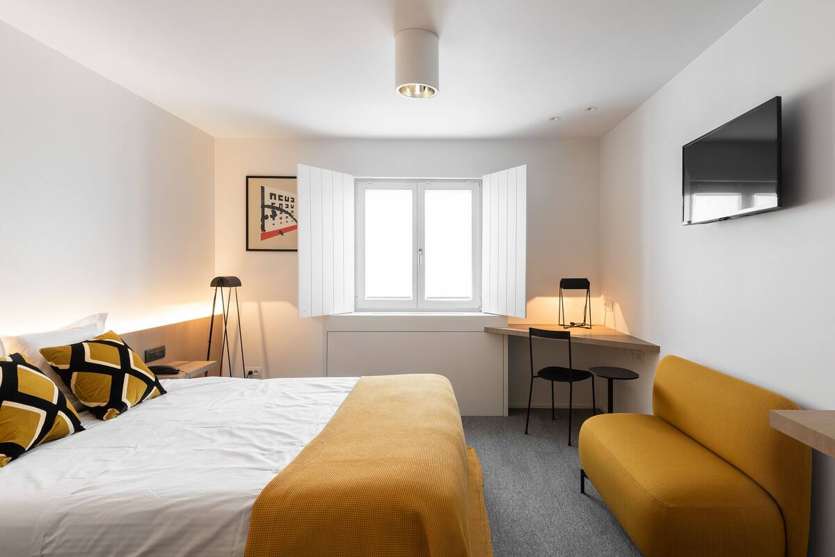 A hotel room with a bed, a desk, a chair, a sofa, a TV, and a window.