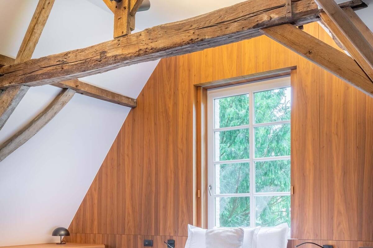 A bedroom with a sloped ceiling, wooden beams, a large window, a bed with white bedding, and a small bedside table.
