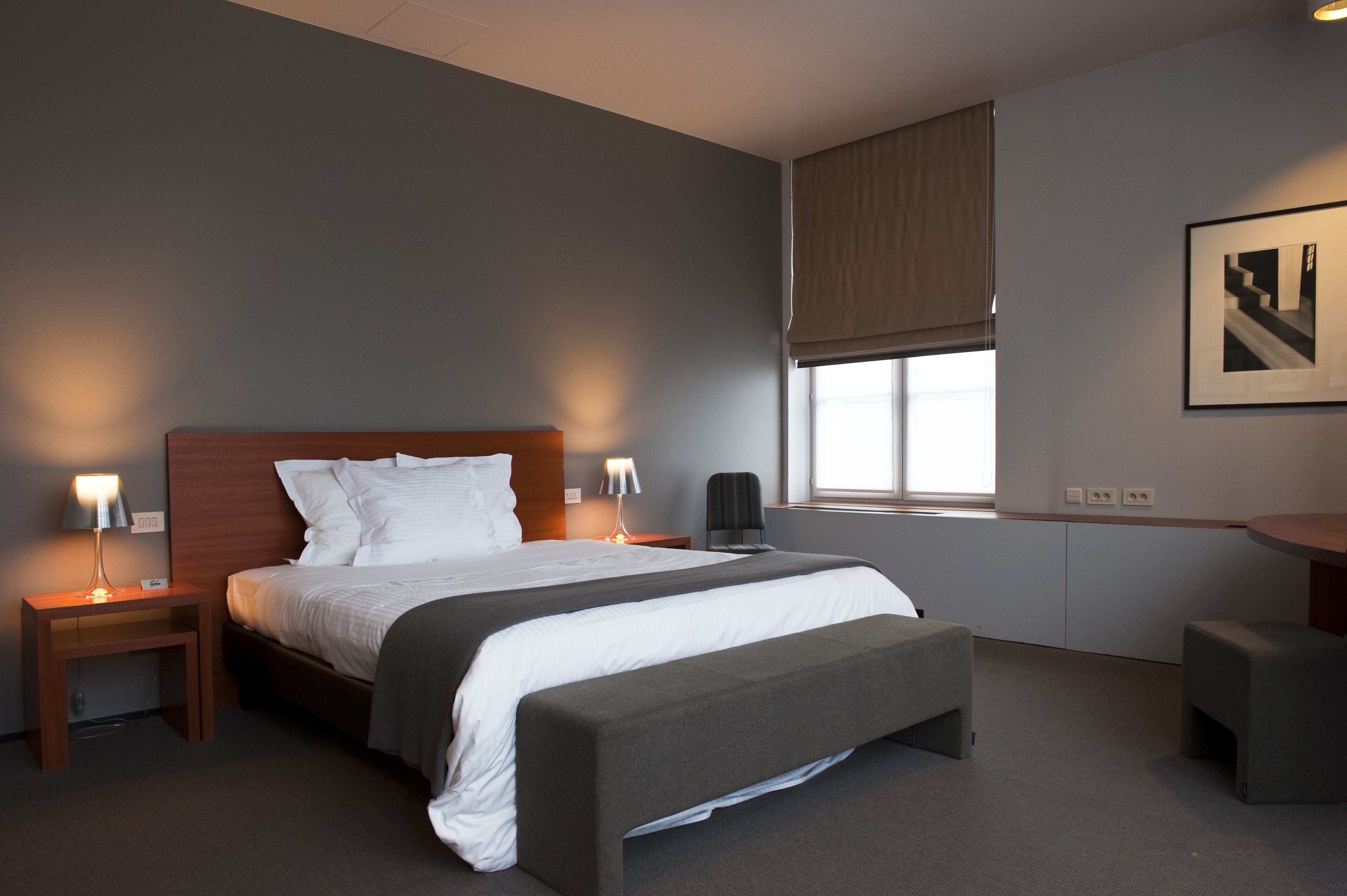 Quarto de hotel moderno com uma cama grande, mesas de cabeceira, um banco, uma escrivaninha e uma janela com persianas.
