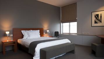 Quarto de hotel moderno com uma cama grande, mesas de cabeceira, um banco, uma escrivaninha e uma janela com persianas.