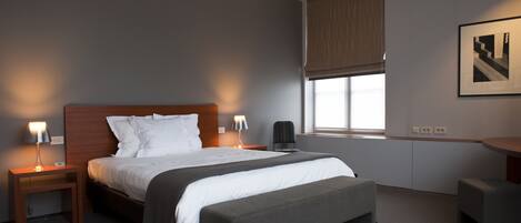 Ein modernes Hotelzimmer mit einem großen Bett, Nachttischen, einer Bank, einem Schreibtisch und einem Fenster mit Jalousien.