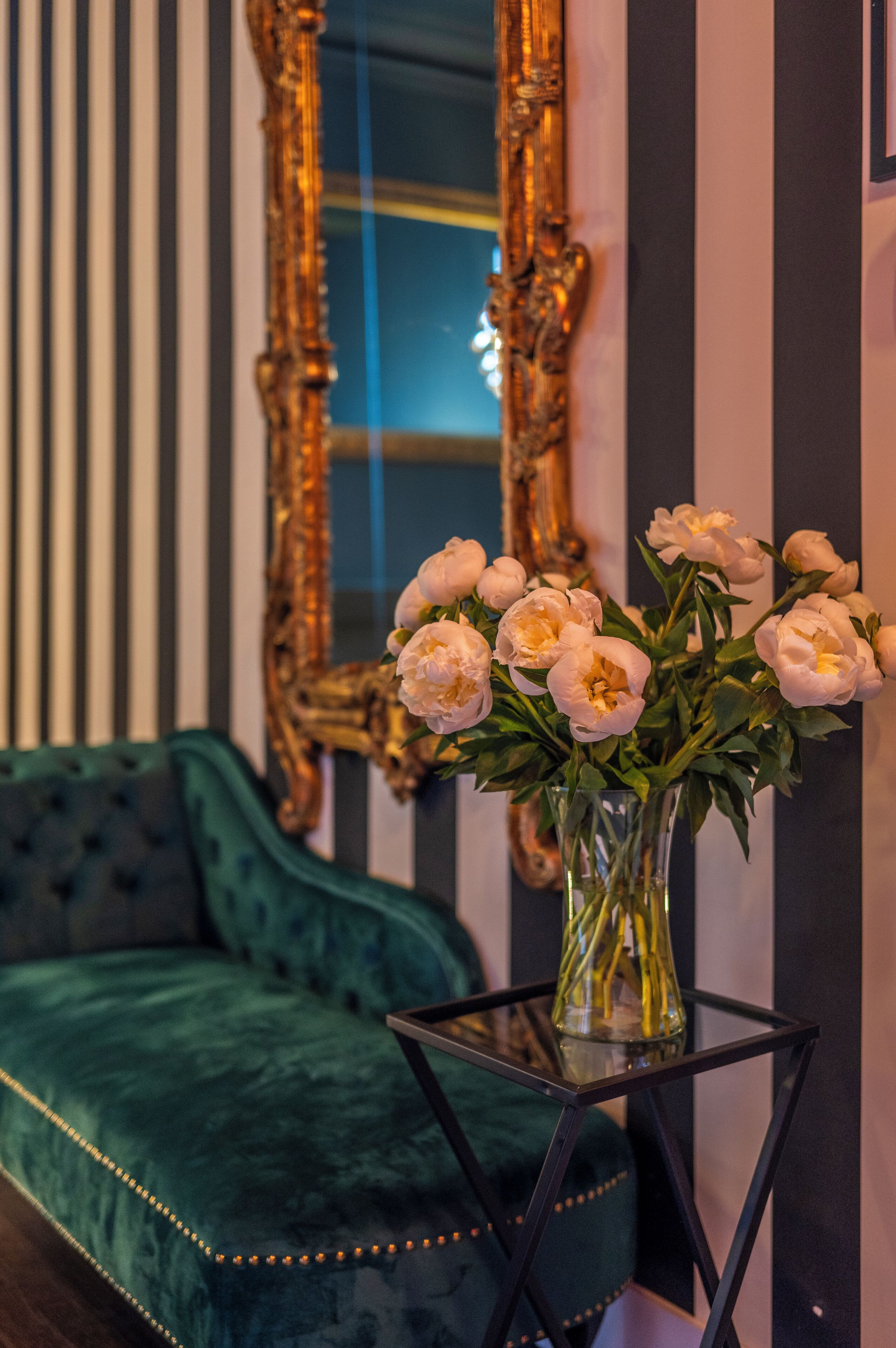 Una habitación con un sofá de terciopelo verde, un jarrón de cristal con flores y un espejo con marco dorado.
