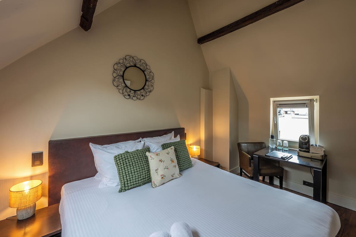 Una cama bien hecha con ropa de cama blanca y almohadas estampadas en verde, una mesita de noche con una lámpara, y un escritorio con una silla y una ventana.