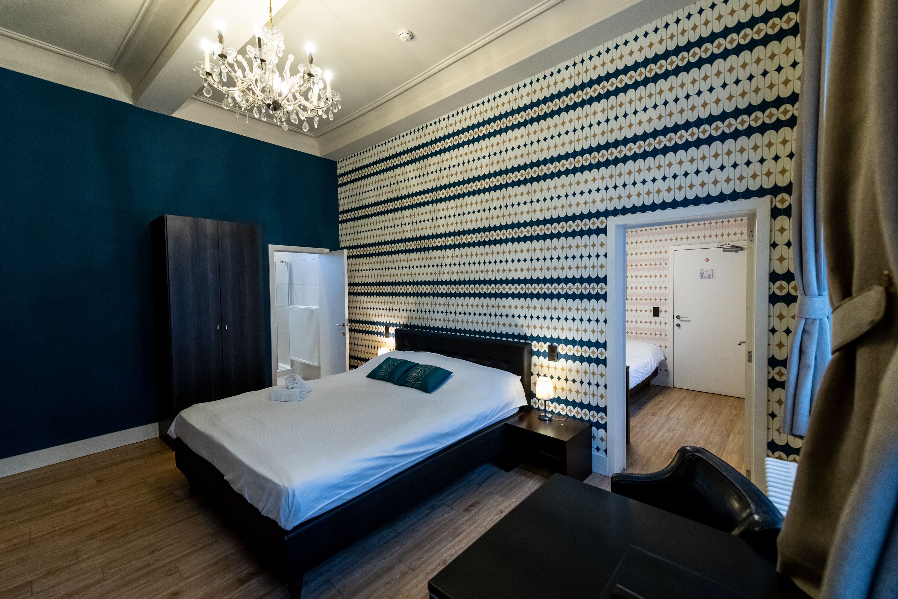 Een slaapkamer met een bed, nachtkastje, dressoir, een kroonluchter en een behang met patroon.