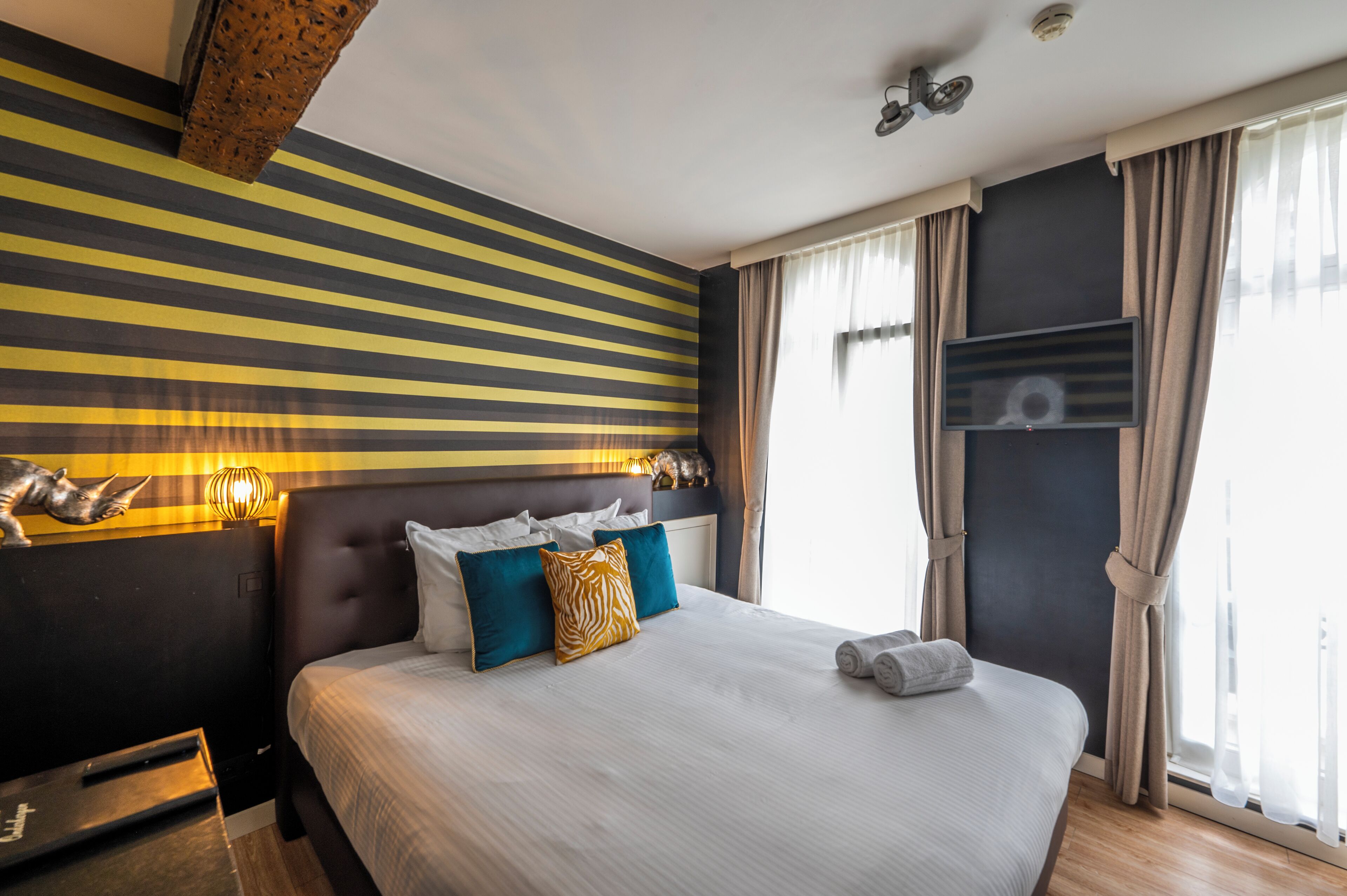 A hotel room with a striped wall, a large bed with multiple pillows, a nightstand, and a window with curtains.