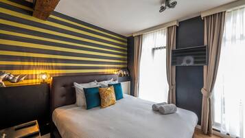 A hotel room with a striped wall, a large bed with multiple pillows, a nightstand, and a window with curtains.
