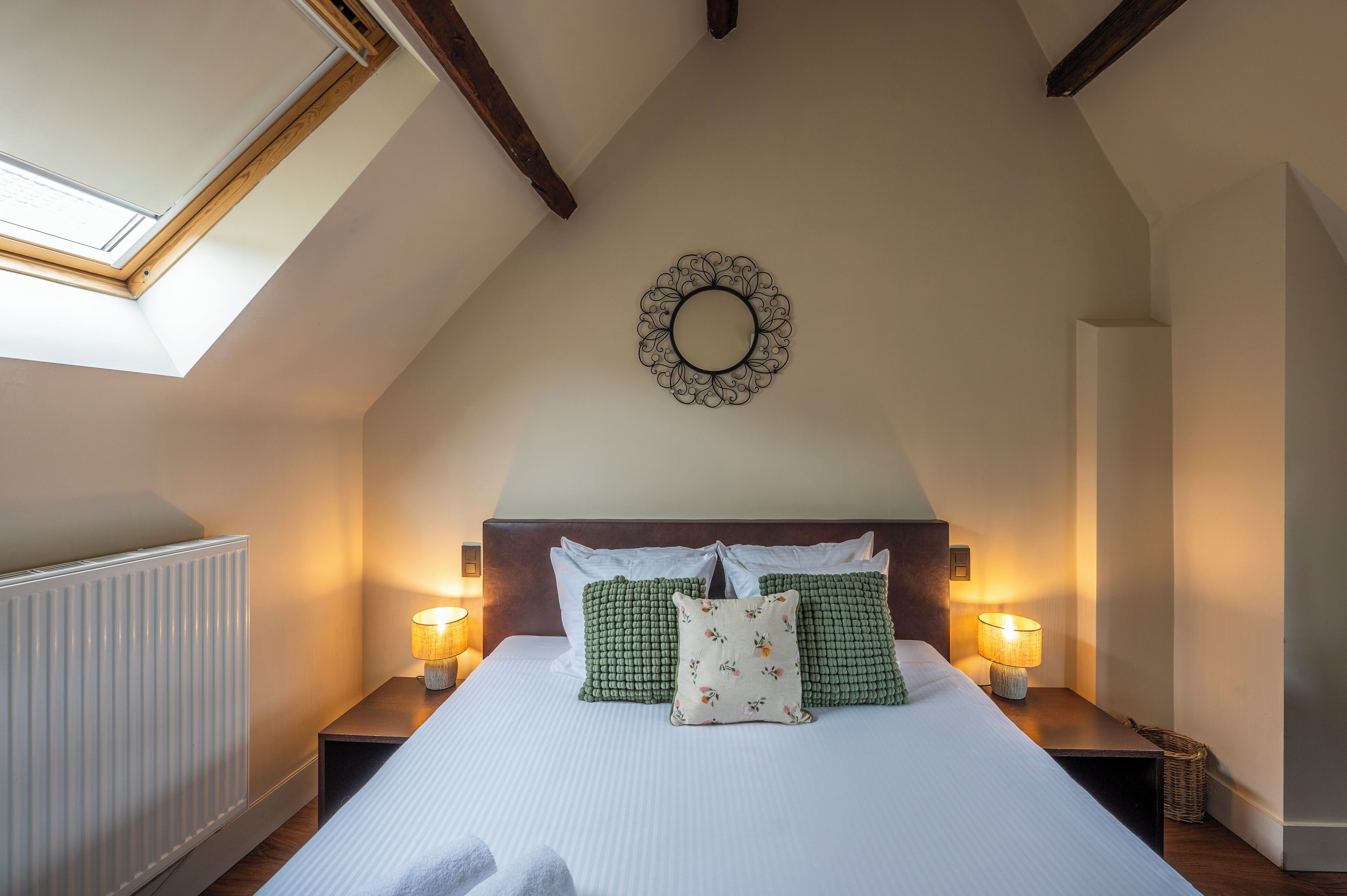 Une chambre avec un plafond en pente, un puits de lumière, un lit avec une literie blanche, des coussins verts et à motifs floraux, deux lampes de chevet et un miroir décoratif sur le mur.